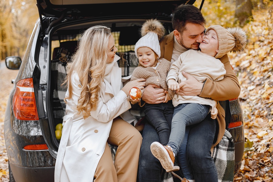 Happy family resting after day spending outdoor in autumn park. Father, mother and two children sitting inside car trunk, smiling. Family holiday and traveling concept.